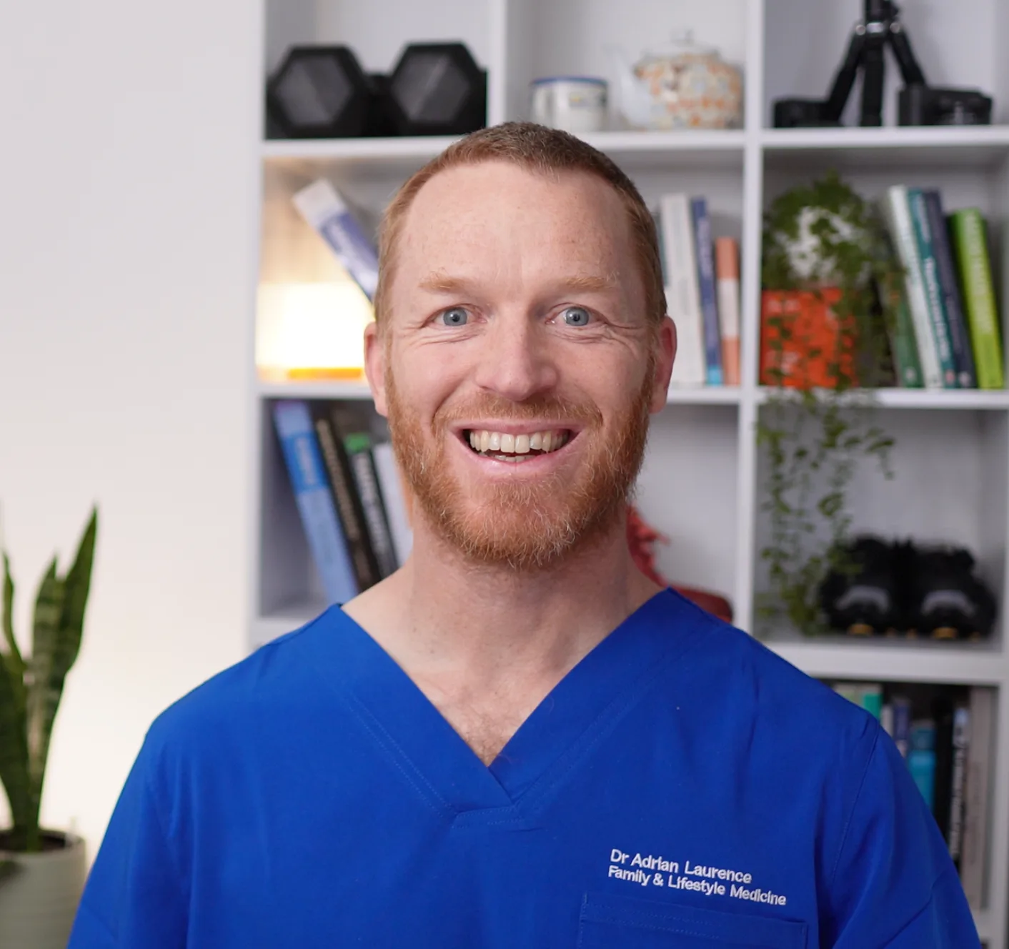 Dr Adrian Laurence, smiling, in blue Family and Lifestyle Medicine scrubs, in front of a bookshelf with medical texts, a plant, and exercise equipment.